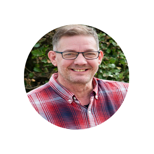 Jonathan Jones in a red plaid shirt with glasses and a smiley face stood in front of a green hedge.